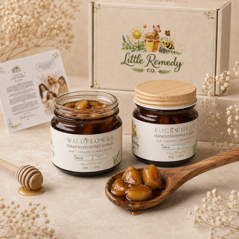Two jars of fermented honey garlic with a wooden spoon filled with honeycomb pieces, set against a 'Little Remedy Co.' branded box.